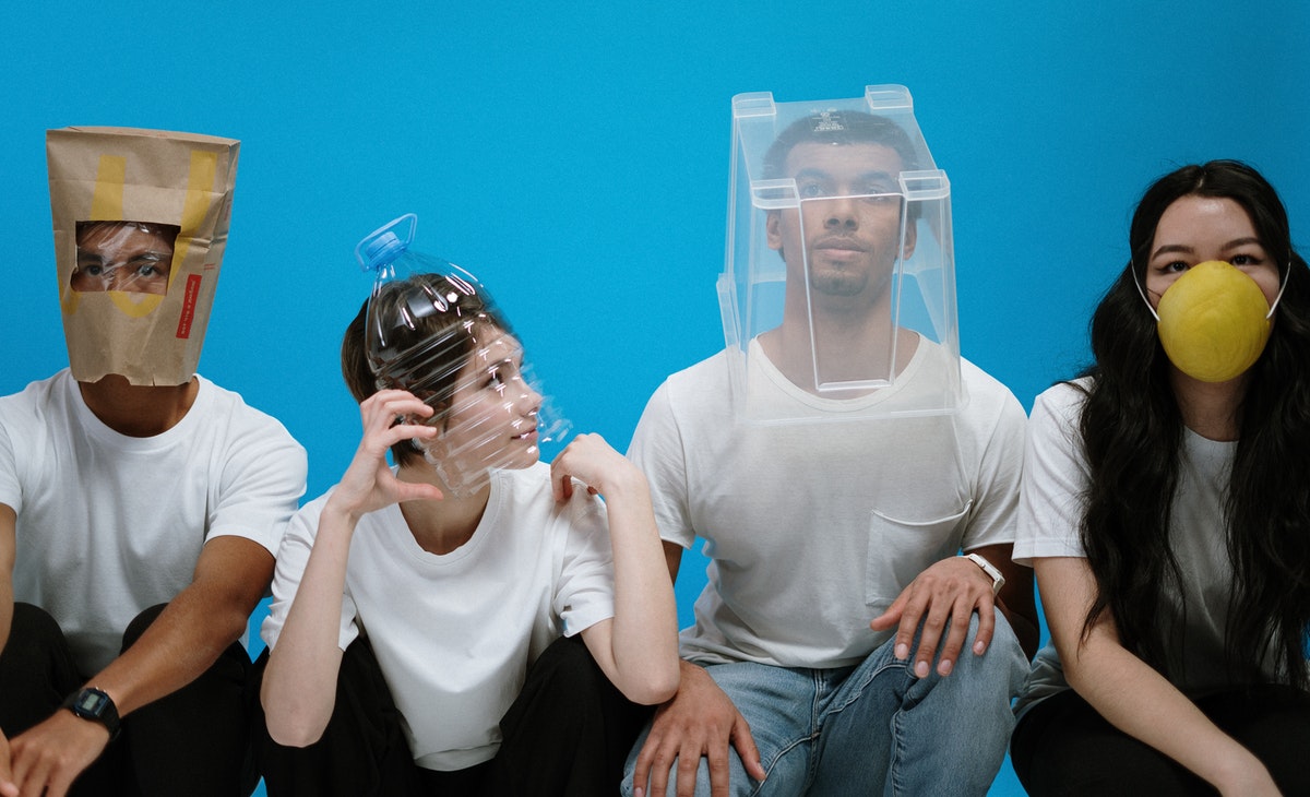 A group of four individuals are seated or crouching against a solid blue background, each wearing a makeshift face covering. From left to right: a person with dark skin wears a brown paper bag over their head with cutouts for eyes and mouth; a person with light skin has the top half of a clear plastic water bottle fitted over their face; a person with dark skin has a clear, rectangular plastic container over their head; and a person with light skin wears a bright yellow, spherical mask over their nose and mouth. All four are wearing white t-shirts, and the two individuals on the left are also wearing dark pants.