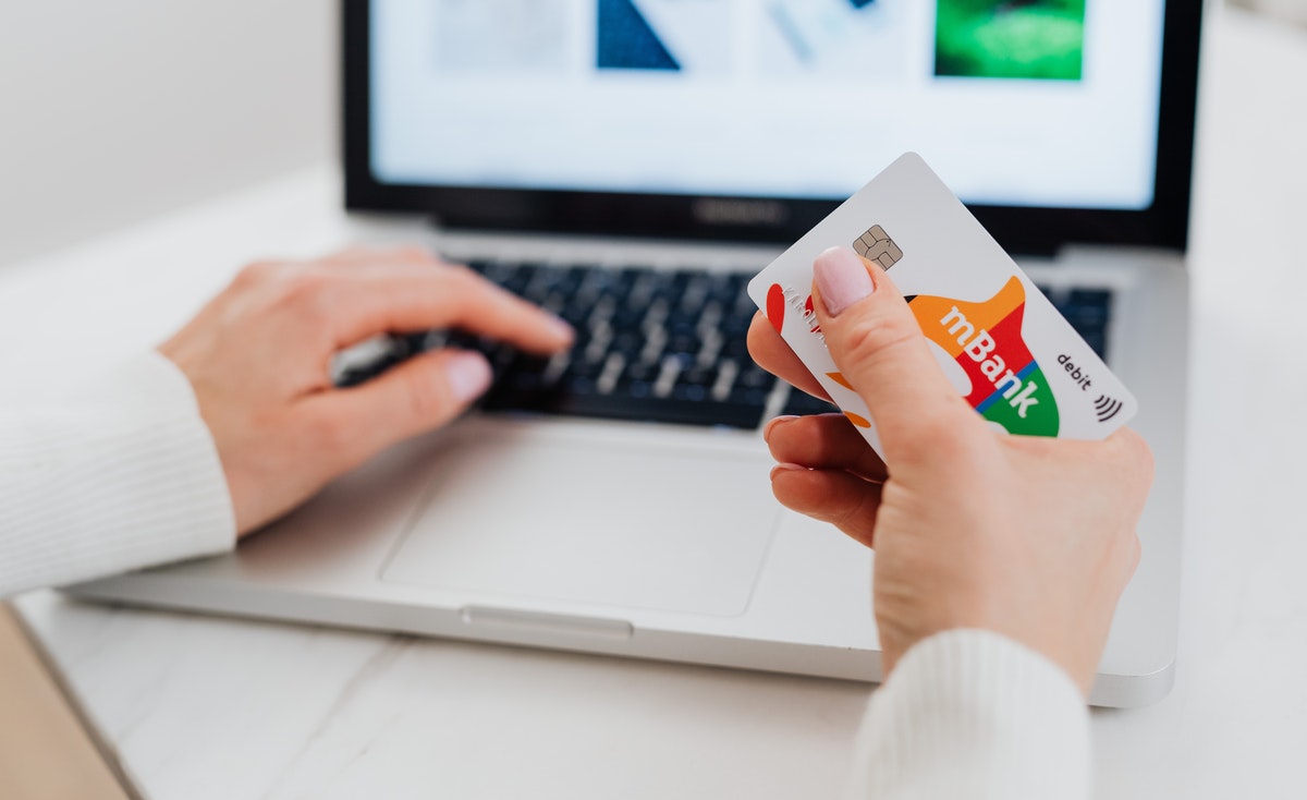 A close-up shot focuses on a person's hands as they engage in online shopping or banking. The person's left hand, with manicured nails, is positioned over the keyboard of an open silver laptop, seemingly typing. Their right hand holds a white "mBank" debit card with colorful letters (orange "m," red "B," green "a," yellow "n," and red "k") and a small signal icon for contactless payment. The laptop screen is blurred in the background but shows what appears to be a website interface. The setting suggests a clean, possibly home or office, environment with a light-colored surface.