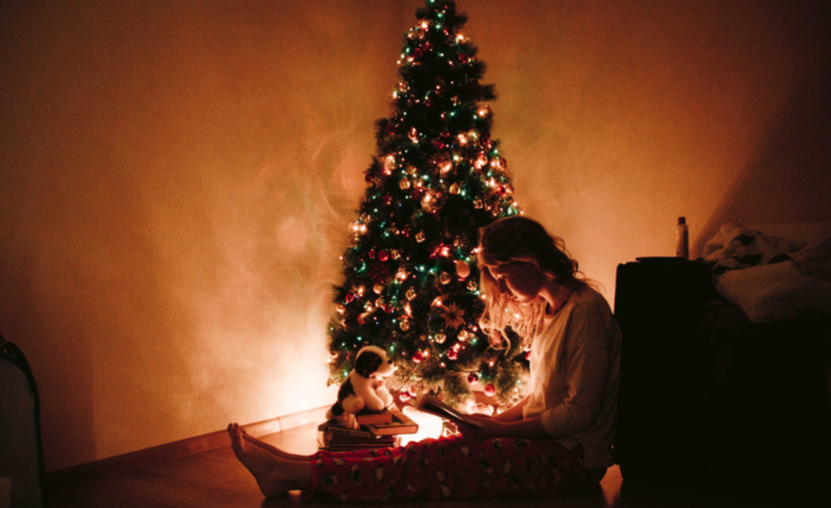 A young woman sits thoughtfully beside a beautifully decorated Christmas tree adorned with lights, baubles, and ornaments. She appears reflective or melancholic, evoking a quiet, emotional moment during the holiday season.