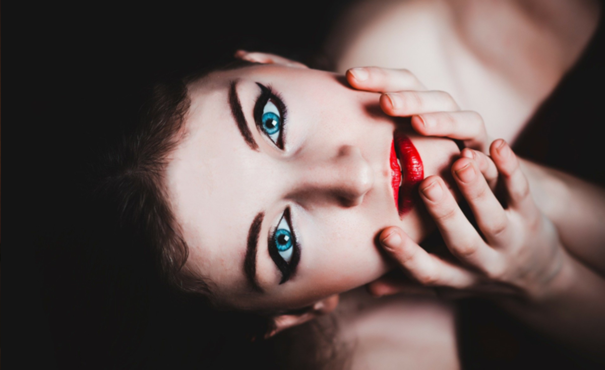 A close-up portrait of a woman's face with her hand covering her mouth, cropped at an angle. Her eyes are expressive, framed by dark eyebrows and eyeliner, and her fingers rest gently over her lips, suggesting surprise or contemplation. The lighting is soft and moody, creating an intimate and thought-provoking mood.