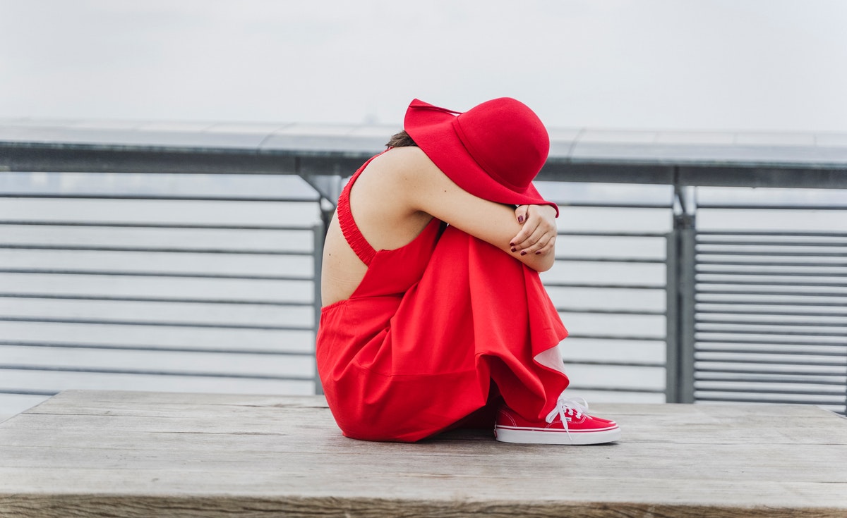 A woman dressed in red sits alone on a wooden platform with her head resting on her knees, arms wrapped around her legs. She appears distressed or emotionally overwhelmed. The setting is urban, with a metal railing and overcast background suggesting isolation or vulnerability in a public space.