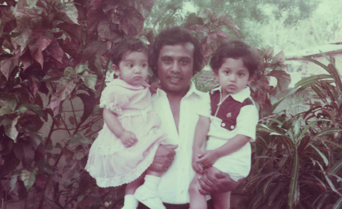 A father standing outdoors holding his two young children, a daughter in a pink dress and a son in a white outfit with maroon accents. They are surrounded by lush tropical plants in what appears to be a garden setting, likely taken in the 1980s.