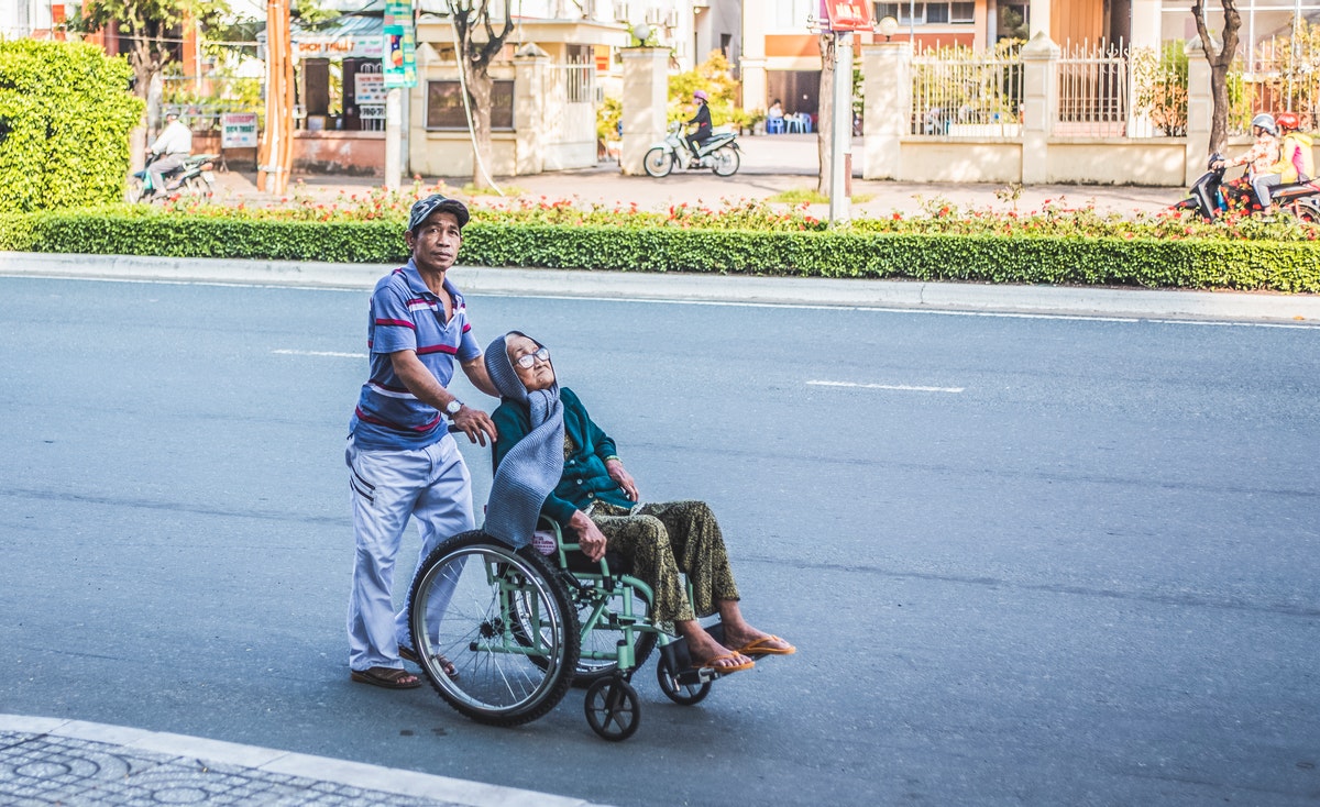 A slightly elevated eye-level shot captures a middle-aged man pushing an elderly woman in a wheelchair on the right side of a paved road. The man, on the left, is wearing a striped short-sleeved shirt, light-colored pants, and a cap, looking slightly upwards and to the left. The woman, on the right, is seated in a light green wheelchair, wearing a dark green top, patterned pants, and a dark scarf wrapped around her head and neck. She looks upwards with a serene expression. The road is clear, with a low, well-maintained hedge and a sidewalk on the right side, separating it from houses and what appear to be power poles and trees. In the background, on the road, a person on a motorcycle is visible. The scene is brightly lit by natural light.