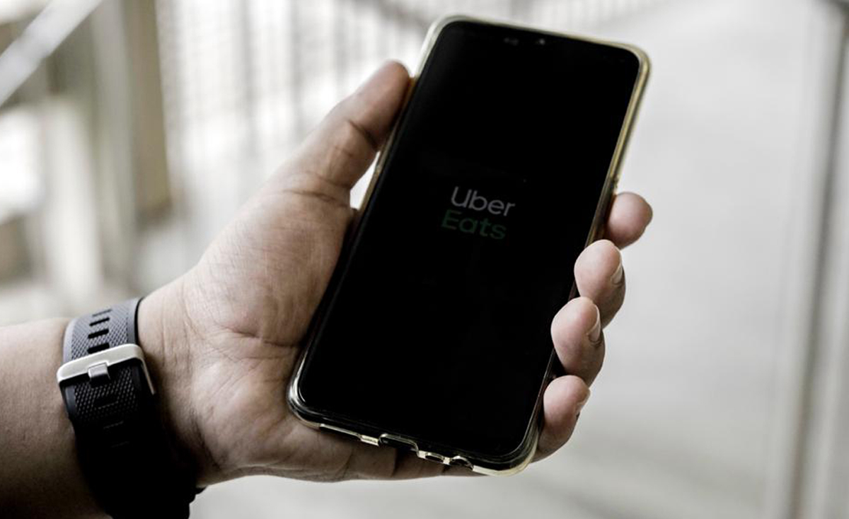A left hand, adorned with a black smartwatch, holds a smartphone displaying the Uber Eats logo in white and green text on a black screen. The phone is encased in a translucent, golden-hued protective case. The background is a blurred, bright indoor setting with light-colored steps or railings.