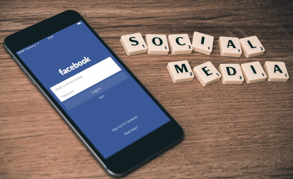 A smartphone displaying the Facebook login page is positioned on a wooden surface next to nine ivory-colored Scrabble-like tiles. The tiles are arranged to spell "SOCIAL MEDIA" with "SOCIAL" in the top row and "MEDIA" below it. The phone's screen shows fields for "Email or phone number" and "Password," along with "Log In" and "Back" buttons.