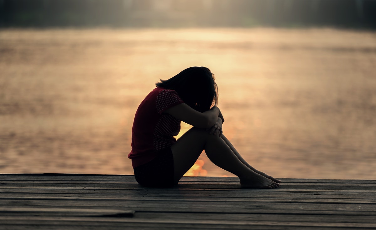 A somber image depicts a person in silhouette, sitting alone on a wooden pier with their head bowed and arms wrapped around their knees, facing a body of water reflecting the setting or rising sun. The scene evokes feelings of profound sadness, isolation, and contemplation of suicide.