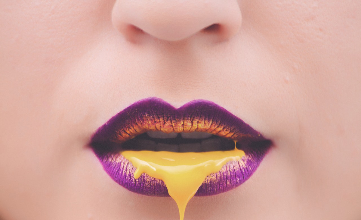 A vibrant close-up shot focuses on a young woman's mouth with striking makeup. The lips are painted a deep purple with golden highlights, and a thick stream of bright yellow liquid drips from the lower lip. The tip of the nose is visible at the top of the frame, and the surrounding skin is smooth. The image has a slightly surreal or artistic quality.
