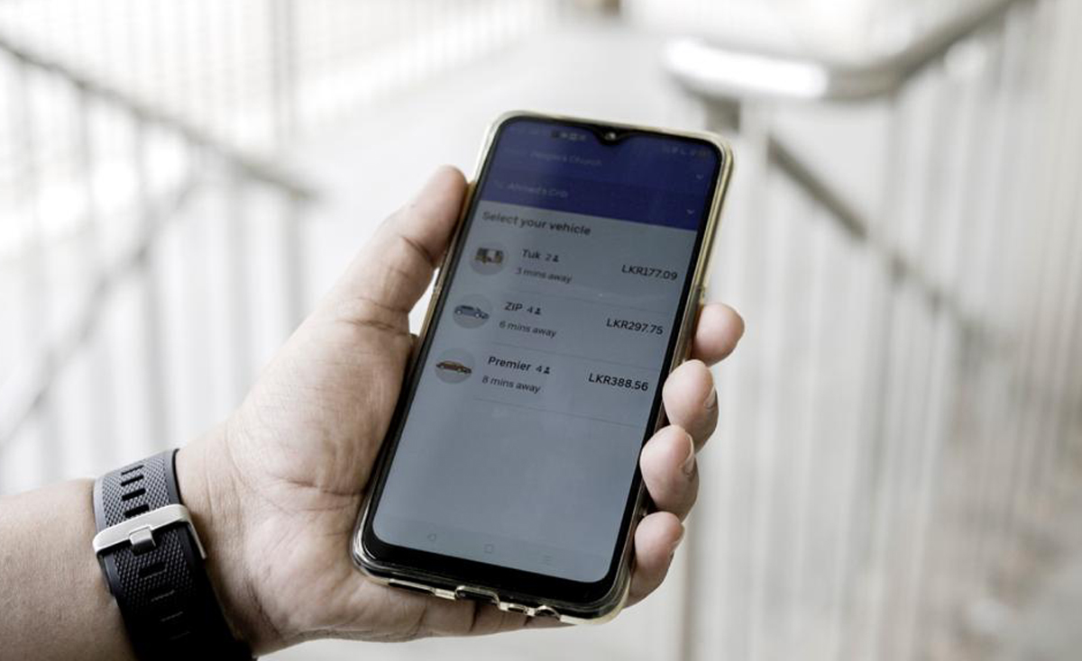 A left hand, wearing a black smartwatch, holds a smartphone displaying a ride-hailing app (Uber) interface. The screen shows options to "Select your vehicle" with categories like "Tuk Tuk," "ZIP," and "Premier," along with estimated prices in LKR (Lankan Rupees) and distance/time estimates. The phone is in a translucent, golden-hued case, and the background is a blurred, bright indoor setting with light-colored steps or railings.