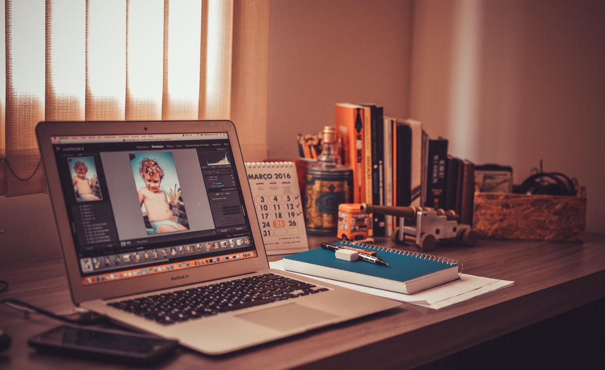 An indoor desk setup for working smart features an open MacBook on the left displaying photo editing software with an image of a child. To the right of the laptop, a teal notebook with a pen rests on a piece of paper, and behind it, a "MARCO 2016" calendar. In the background, a collection of books, decorative items, and a wicker basket are visible, all bathed in warm, soft light from a nearby window with vertical blinds, suggesting a productive and organized workspace.