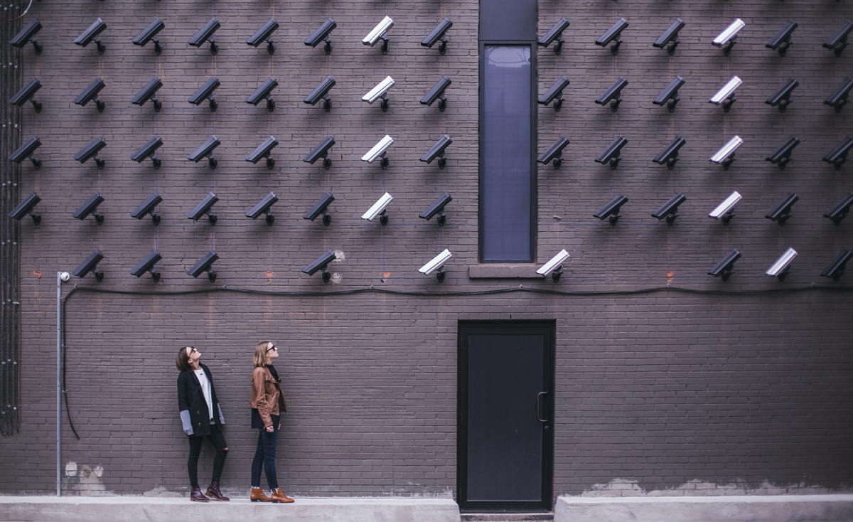 Two women stand on a sidewalk against a large brick wall, looking up at the dozens of security cameras mounted in neat rows across the wall above them.