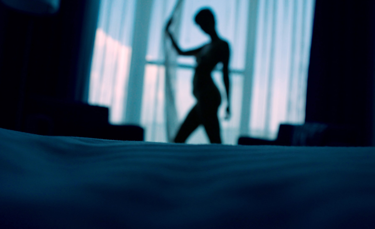 A low-angle view from across a bed showing the silhouette of a woman's figure standing in front of a bright window. She is holding a sheer curtain in a dimly lit, blue-toned room.