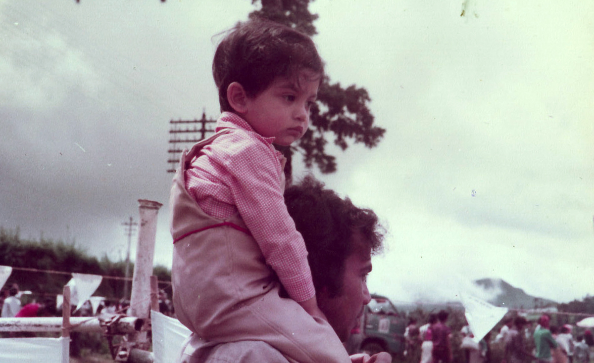 A slightly faded, low-angle shot shows a young child with dark hair sitting on the shoulders of an adult, who is partially visible on the right. The child, wearing a light-colored long-sleeved shirt and pants, looks thoughtfully to the right. In the background, there are trees, a cloudy sky, and faint indications of a gathering with white flags or banners, suggesting an outdoor event like a road race.