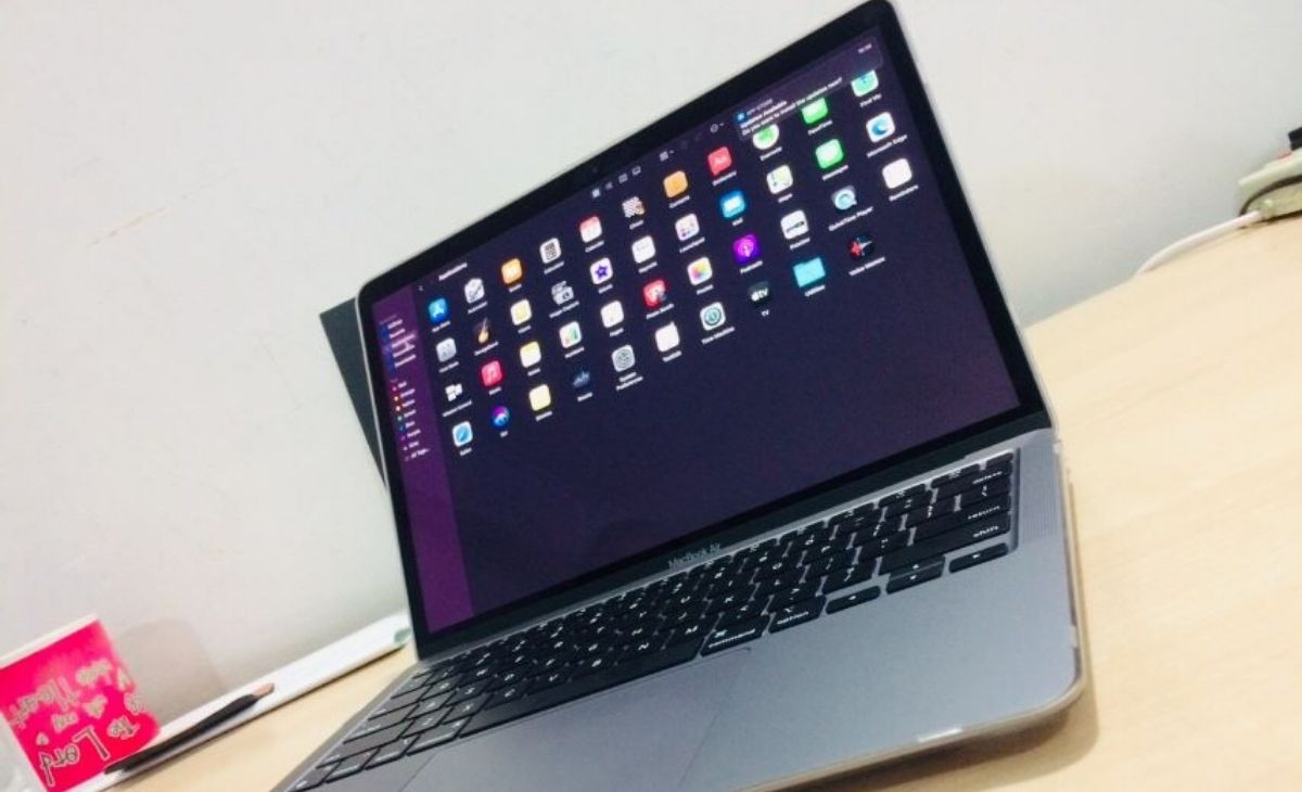 A close-up, high-angle shot captures an open Space Gray MacBook Air laptop placed on a light wooden desk, angled from the lower right towards the upper left. The screen is illuminated, displaying a dark background with numerous colorful application icons arranged in a grid. The keyboard is visible, showing the black keys. To the left of the laptop, a red mug with white text and a dark-colored pen are partially visible. The background consists of a plain white wall, with a hint of a power outlet or similar fixture in the upper right corner.
