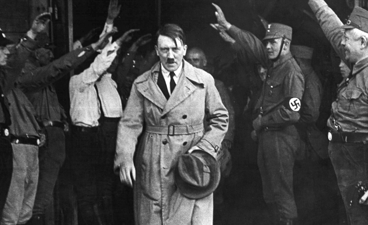 A black and white historical photo of Adolf Hitler in a trench coat and tie, walking forward while surrounded by men in uniform giving the Nazi salute. Hitler is holding a hat in his left hand.