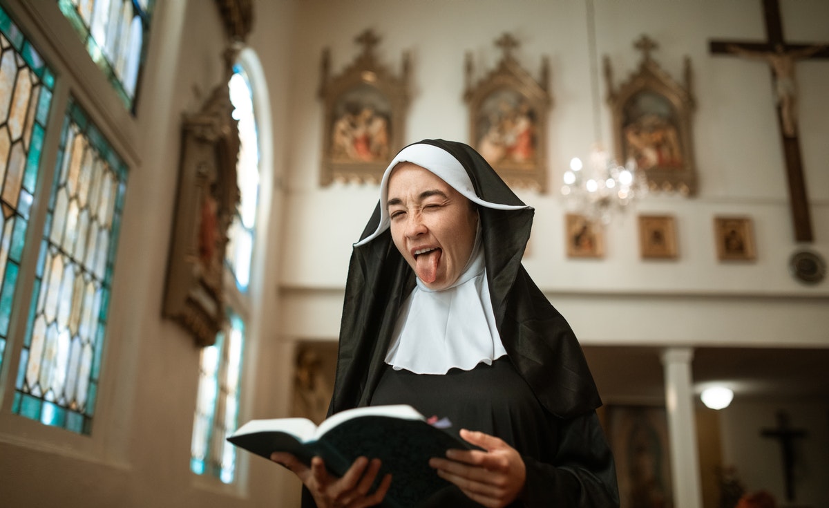 A medium eye-level shot captures a nun in a black habit and white wimple, standing in what appears to be a church interior. She is holding an open book with both hands and has a humorous or defiant expression, sticking her tongue out and squinting her eyes. In the background, stained-glass windows are visible on the left, and religious artwork depicting scenes from the Stations of the Cross, along with a crucifix and a chandelier, adorn the far wall. The lighting is bright, illuminating the nun and the church's interior.