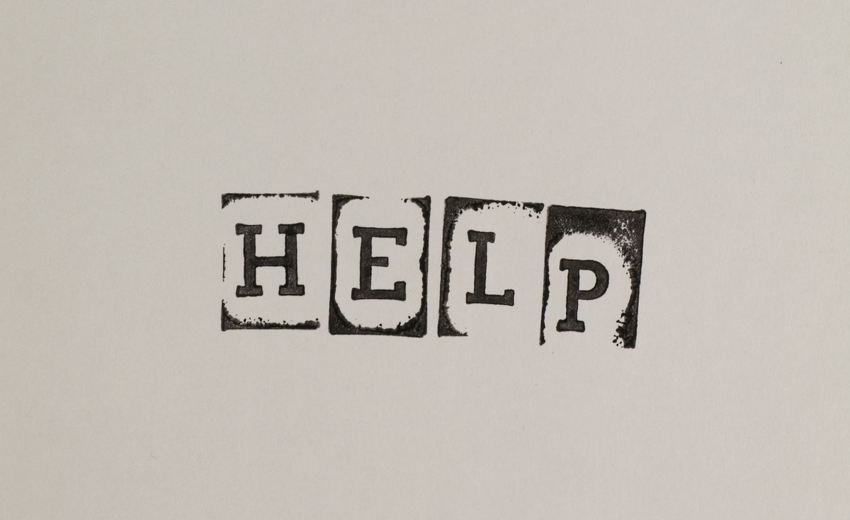 A close-up, eye-level shot captures the word "HELP" typed or stamped in bold, blocky, distressed black letters on a plain, light grey or off-white background. Each letter appears to be individually stamped, with slightly uneven edges and some ink smudges, particularly around the "P". The spacing between the letters is consistent.