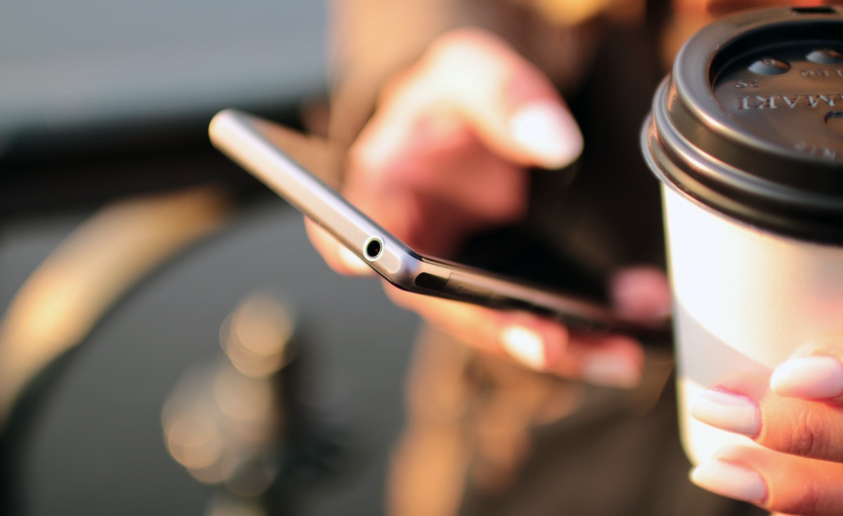 A close-up shot focuses on a person's hands holding a black smartphone and a disposable coffee cup. The right hand is visible at the top, with fingers positioned over the phone's screen, possibly typing or swiping. The left hand holds a white paper coffee cup with a black lid, resting at the bottom of the frame. The smartphone is held at an angle, revealing its side profile with a headphone jack. The background is blurred, showing soft light and dark shapes.
