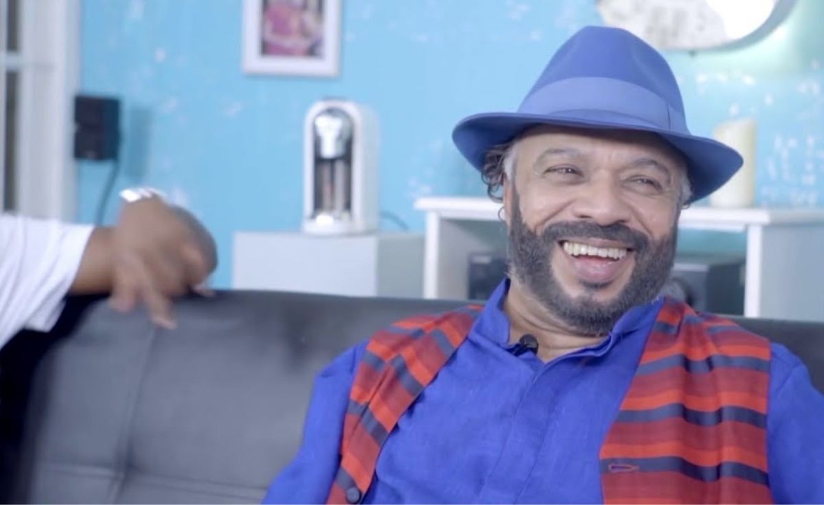 Alt Image Description: A medium eye-level shot features a smiling, middle-aged Sunil Perera with a beard and curly hair, wearing a blue fedora, a blue shirt, and a red and black striped vest. He is seated on a dark leather couch, looking directly at the camera with a wide, joyful smile. To his left, another person's arm is visible, gesturing. The background is a bright blue wall with a framed picture, a white coffee machine, and white shelves. The scene is well-lit.