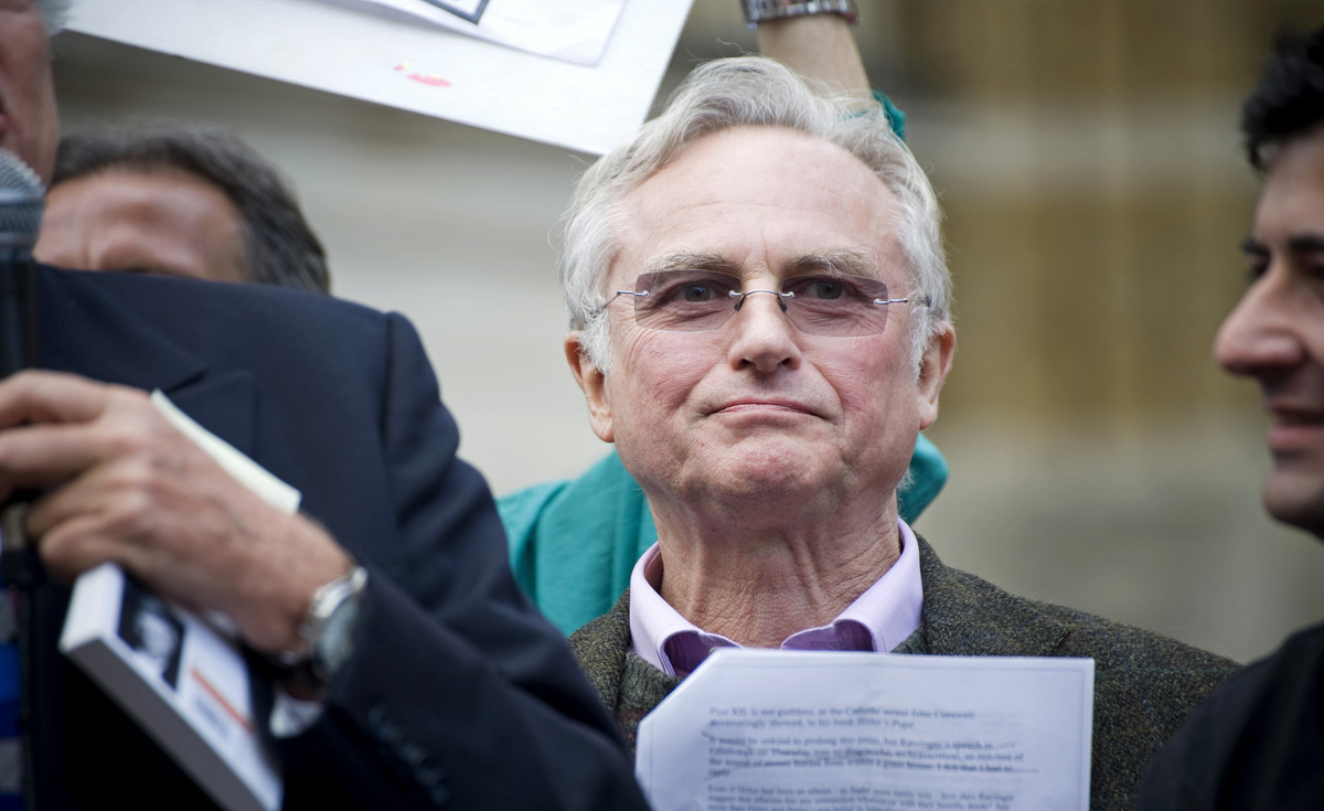 A medium close-up shot features Richard Dawkins, an older man with light grey hair and eyeglasses, looking slightly to his left with a serious expression. He is wearing a light purple collared shirt and a textured dark jacket. To his left, the back of another man's head and shoulder are visible, holding a microphone. To his right, part of another man's face in profile is seen. The background is blurred, suggesting an outdoor or public setting, with faint indications of other people and signs.