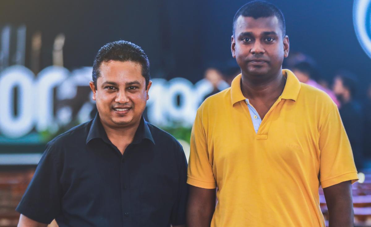A medium eye-level shot features two men standing side-by-side against a dimly lit, blurred background with some faint lettering. The man on the left has light skin, dark hair neatly combed back, and is wearing a dark, possibly black, collared shirt. He is smiling warmly at the camera. The man on the right has darker skin, short dark hair, and is wearing a yellow polo shirt with an unbuttoned collar. He has a more neutral expression, looking directly at the camera. The lighting highlights their faces and upper torsos.