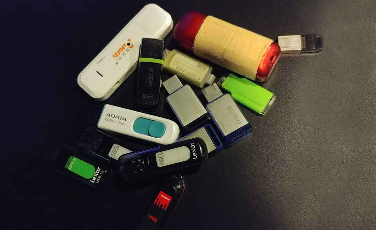 A top-down, close-up shot shows a scattered collection of various USB flash drives on a dark, possibly black, surface. The drives vary in color, size, and design, including white, black, green, silver, and one with a red and beige casing resembling a cork. Some visible brands include "ADATA," "Logiq," "TDK," and "Lexar," with some indicating storage capacities like "4GB," "8GB," and "16GB." The lighting is somewhat dim, creating soft reflections on some of the metallic surfaces.