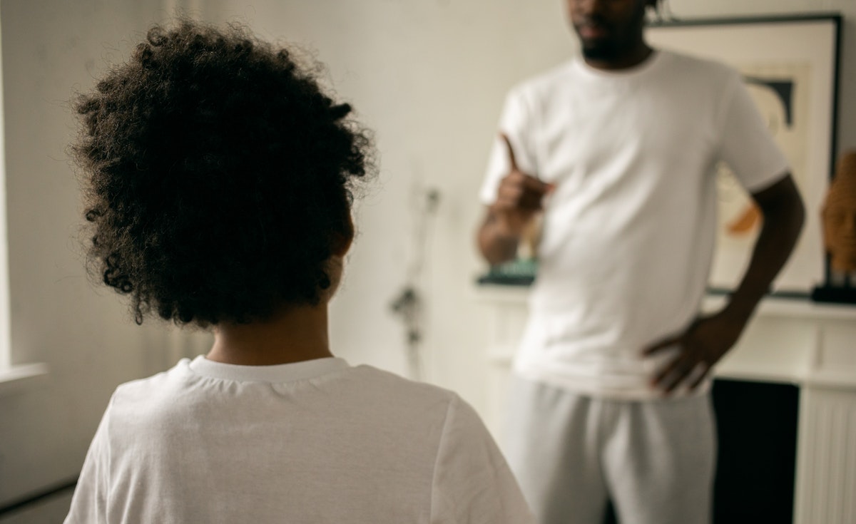 A shallow depth of field shot focuses on the back of a child's head, showing their curly dark hair and the upper portion of their white t-shirt. The child is facing away from the camera. In the blurred background, an adult male, wearing a white t-shirt and light-colored sweatpants, stands with one hand on his hip and the other hand raised with an index finger pointed, as if scolding or instructing. The setting appears to be an indoor room with light walls and a framed picture on the right.
