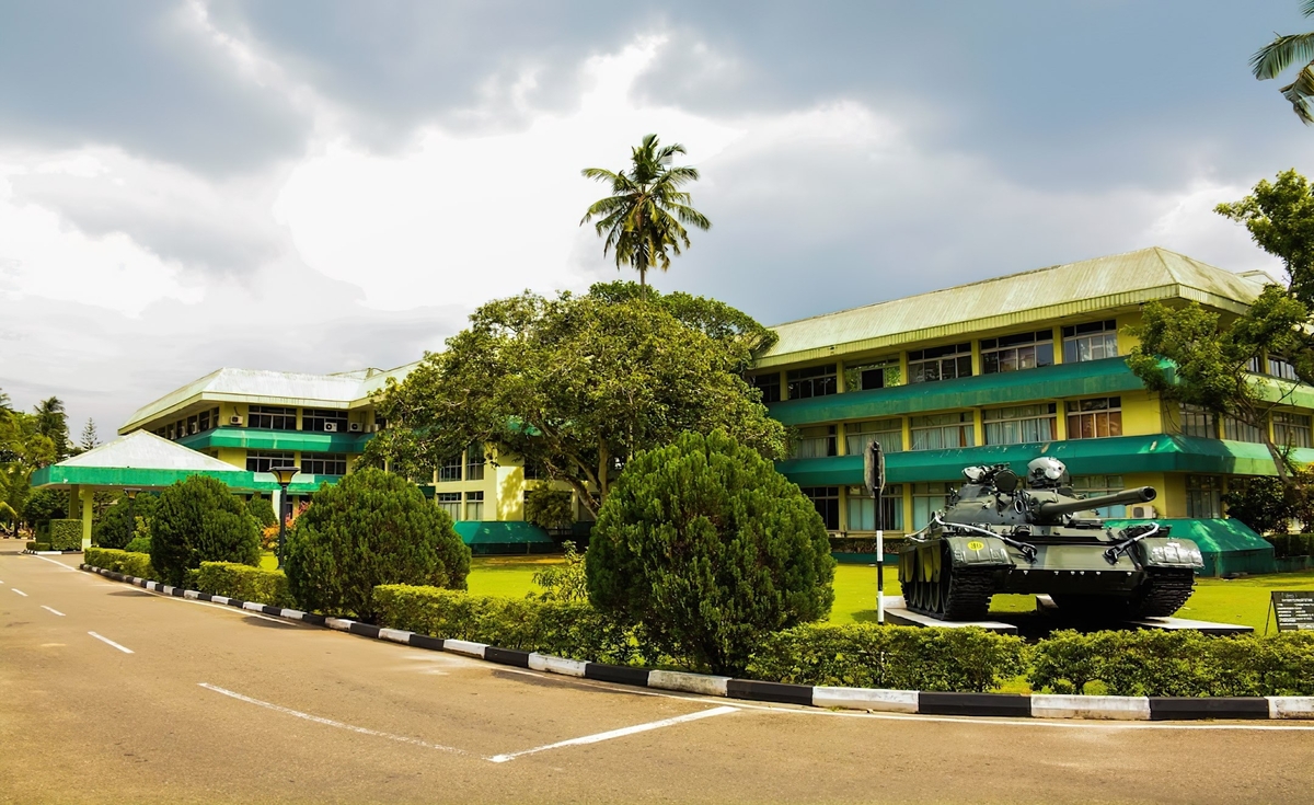 An outdoor, eye-level shot captures a large, multi-story building with a light green roof and balconies, a part of General Sir John Kotelawala Defence University, under a cloudy sky. In front of the building, a dark green tank is displayed on a patch of grass. A paved road with a black and white striped curb curves in the foreground, leading towards the building. Lush green trees and bushes are neatly arranged along the roadside and around the tank, and a tall palm tree rises behind the building.