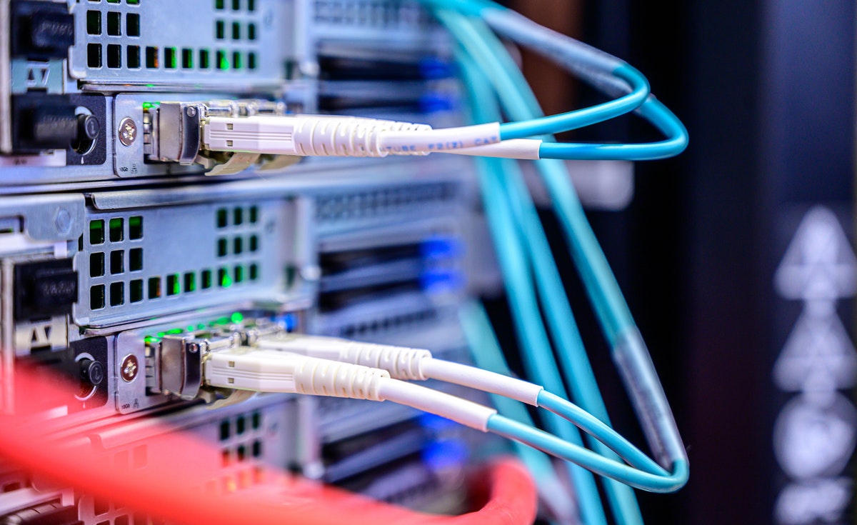 A close-up, eye-level shot focuses on a rack of network servers, densely packed with various cables. Multiple white and blue network cables, likely fiber optic, are plugged into ports on the server units, with their green indicator lights glowing. Some red cables are visible in the foreground, out of focus. The silver server casings and their ventilation grilles are clearly visible, indicating a complex data center or server room environment.