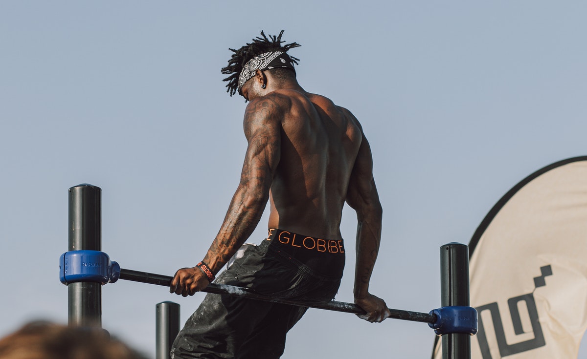 A powerful shot from a slightly low angle captures a muscular, shirtless man with dreadlocks and a black and white bandana, performing an exercise on parallel bars. He is seen from the back, leaning forward with his arms engaged, showcasing his well-defined back and arm muscles. He wears black athletic pants with an orange waistband visible. The sky is bright and clear in the background. A partial view of a white flag with a black symbol is on the right, and a blue base of the exercise equipment is on the left.