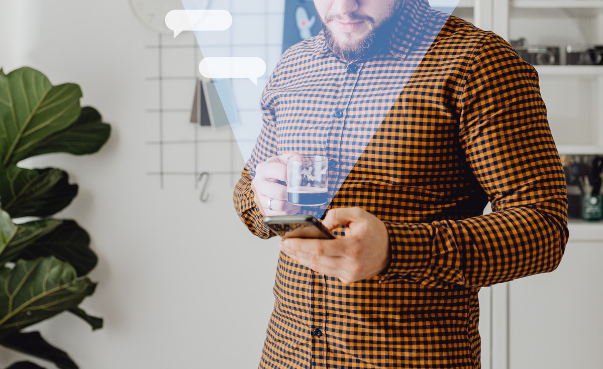 A medium eye-level shot captures a man with a beard, wearing an orange and black plaid shirt, looking down at a smartphone in his left hand. His right hand holds a clear glass mug containing a dark beverage. Overlayed on the image are two white, transparent speech bubbles, suggesting a communication or messaging theme. To the left, a large green plant is partially visible, and the background shows a blurred white wall with shelves and some decorative items.