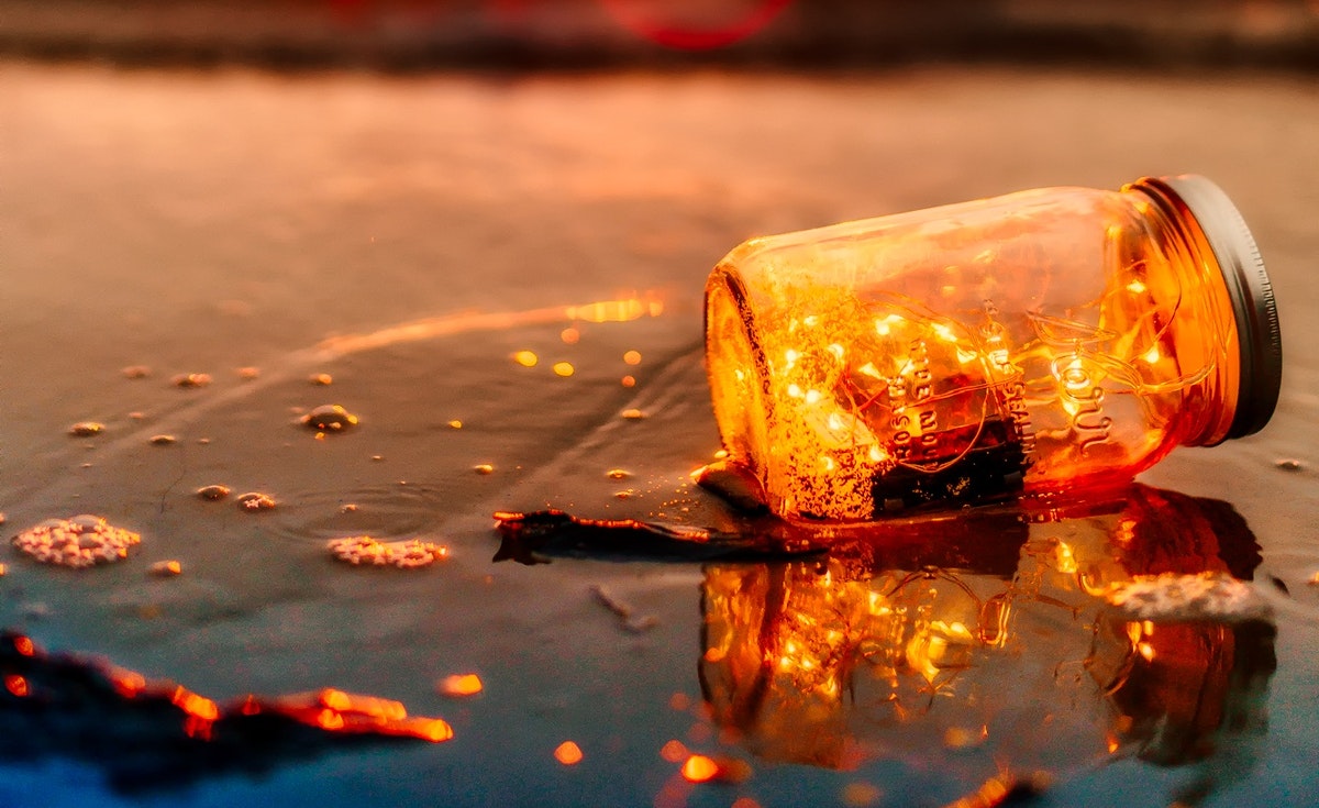 A close-up, horizontal shot captures a clear glass mason jar filled with warm, glowing string lights, lying on its side on a wet, dark surface that reflects the light. The light from the jar casts a bright orange and yellow glow, illuminating the water puddles and creating sparkling reflections. The background is a blurred wash of warm orange and red tones, suggesting a sunset or artificial lighting.