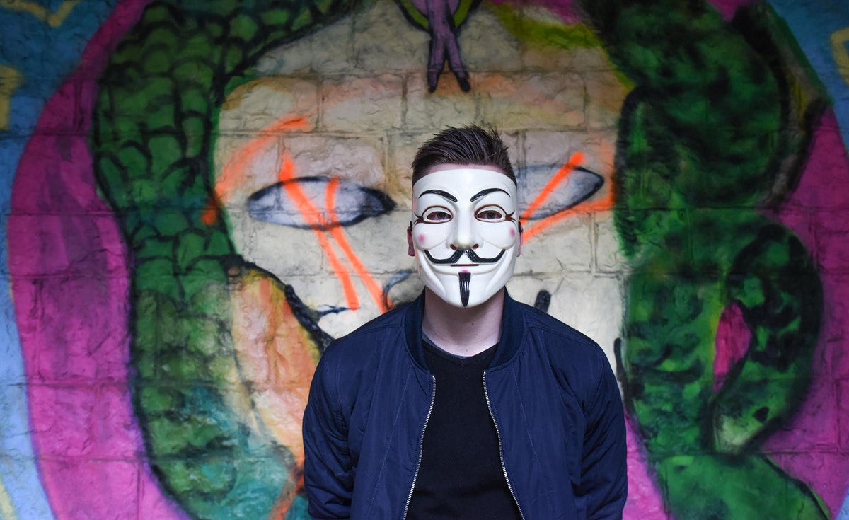 A person wearing a Guy Fawkes mask (also known as a V for Vendetta mask) stands facing forward, looking directly at the camera. They are wearing a dark blue bomber jacket over a black shirt. Behind them is a brightly colored, somewhat abstract graffiti mural depicting a stylized face with snakes or tendrils emanating from it, rendered in shades of green, yellow, pink, and orange. The background is a wall that appears to be made of rough stone or concrete blocks.