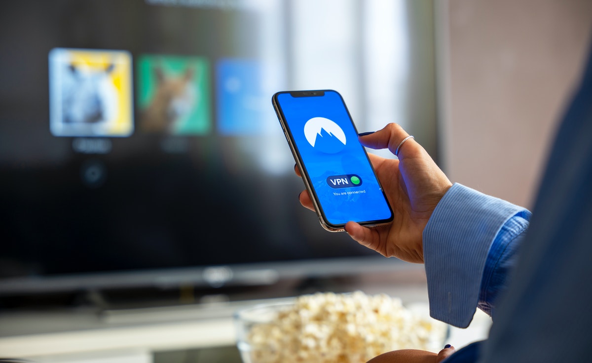 A close-up shot of a person holding a smartphone vertically in their right hand. The phone screen displays a blue interface with a white mountain icon and the word "VPN" below it, along with "You are connected" in white text, indicating an active VPN service. In the blurred background, a television screen shows blurred images, and a bowl of popcorn is visible in the lower foreground. The person's right arm, wearing a light blue striped sleeve, is visible on the right side of the frame.