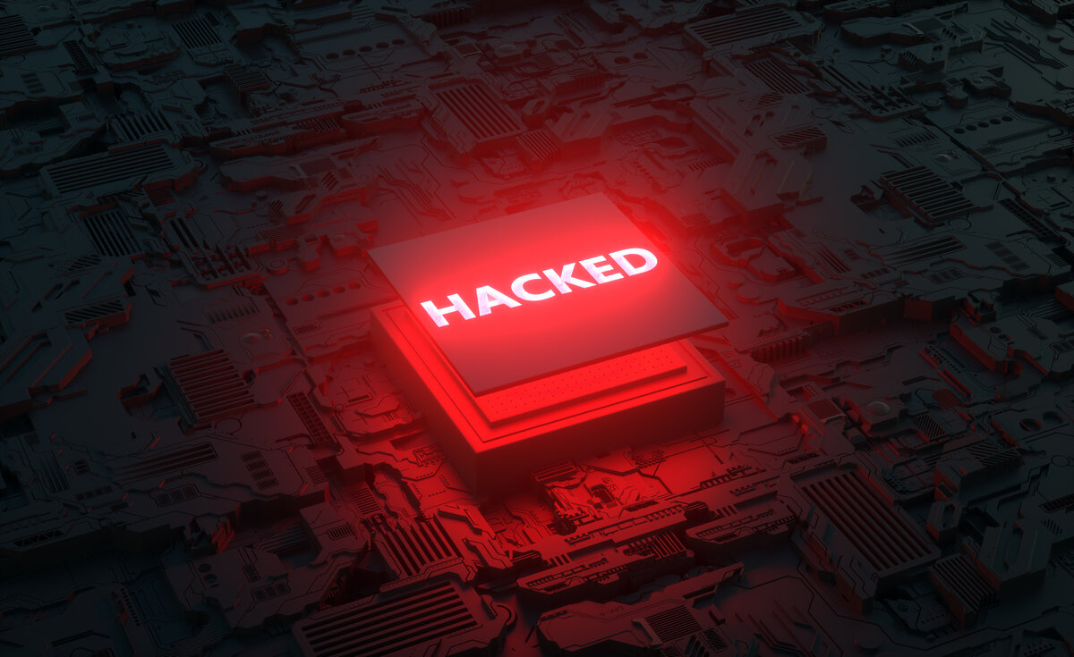 A high-angle, close-up shot captures a glowing red square module, likely a computer chip or core, with the word "HACKED" illuminated in bold white letters across its top surface. This module rests on a dark, intricate circuit board or futuristic cityscape, which is also bathed in a deep red glow from below, creating a sense of urgency and alarm. The detailed textures of the board's components are visible, though somewhat obscured by the strong red light.