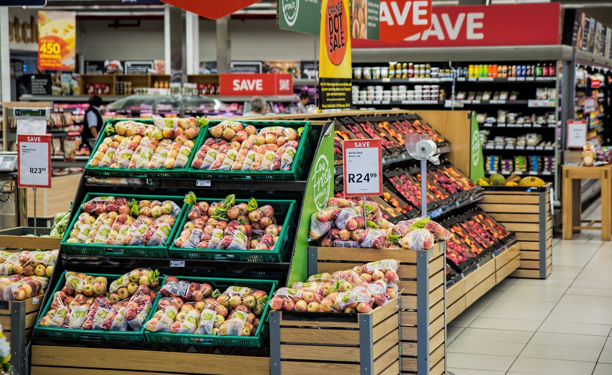 A vibrant supermarket aisle with large displays of fresh produce, including peaches and apples, arranged in green crates and wooden bins. 'SAVE' signs are visible throughout the section.