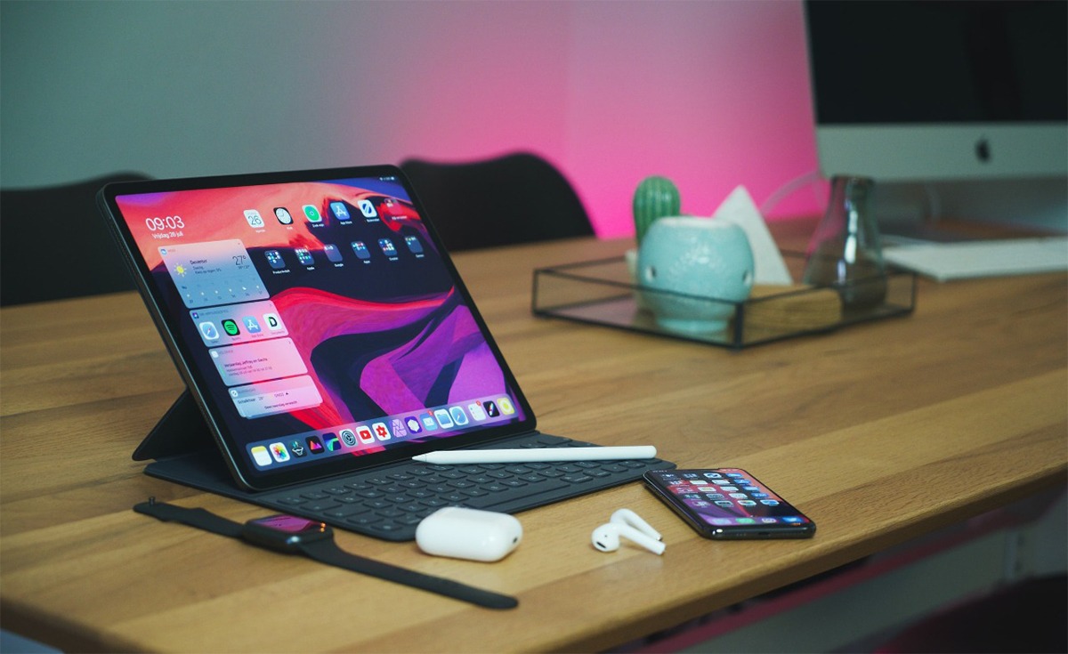 A collection of Apple smart devices on a wooden desk, including an iPad Pro on a keyboard case with a white Apple Pencil beside it, a black Apple Watch, white AirPods in their case, and an iPhone, all against a background with a pink light.