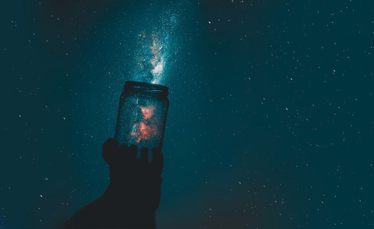 A silhouetted hand holds up a glass jar against a dark, star-filled night sky. A bright, glowing light, resembling a galaxy or nebula, emanates from inside the jar and extends upwards into the sky.