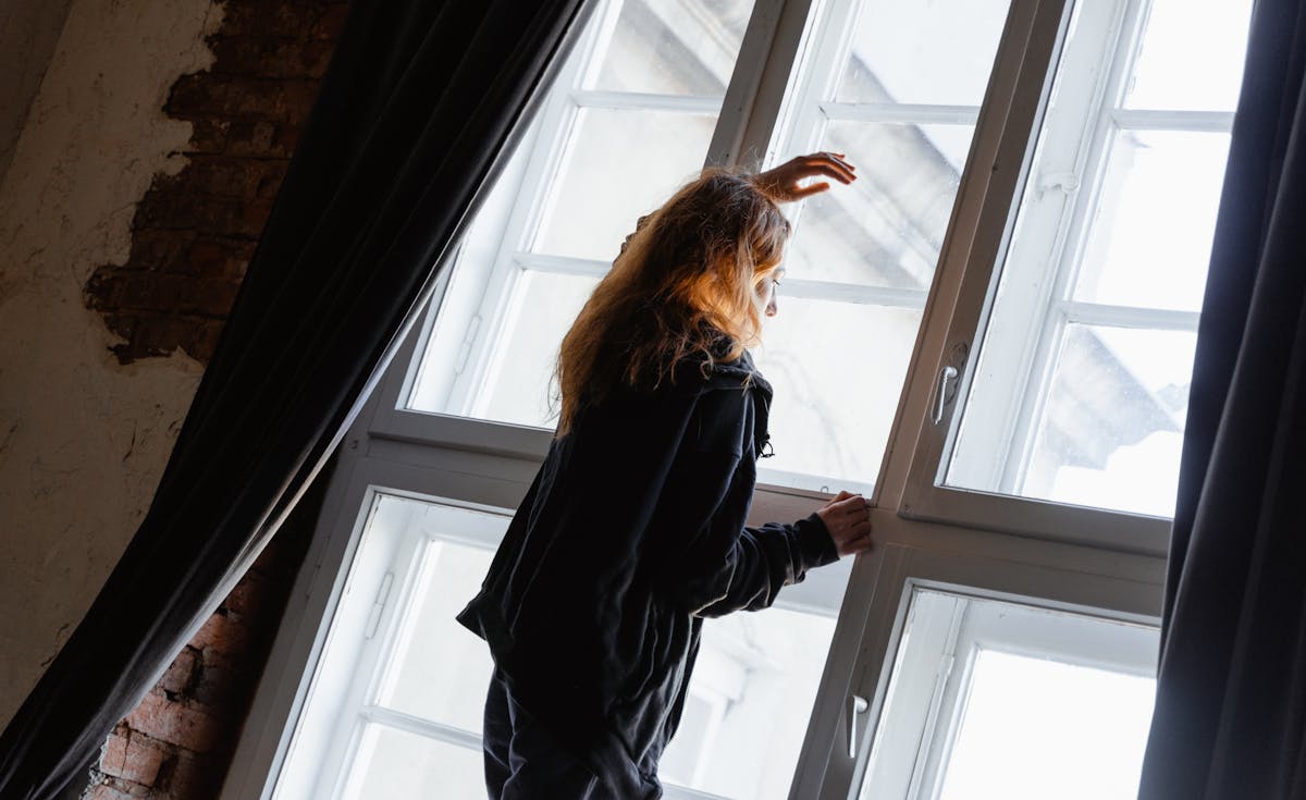 A person with long, wavy brown hair, seen from behind and slightly to the side, stands by a large multi-pane window, reaching up to touch the frame. Dark curtains flank the window, and a hint of a brick wall is visible on the left.