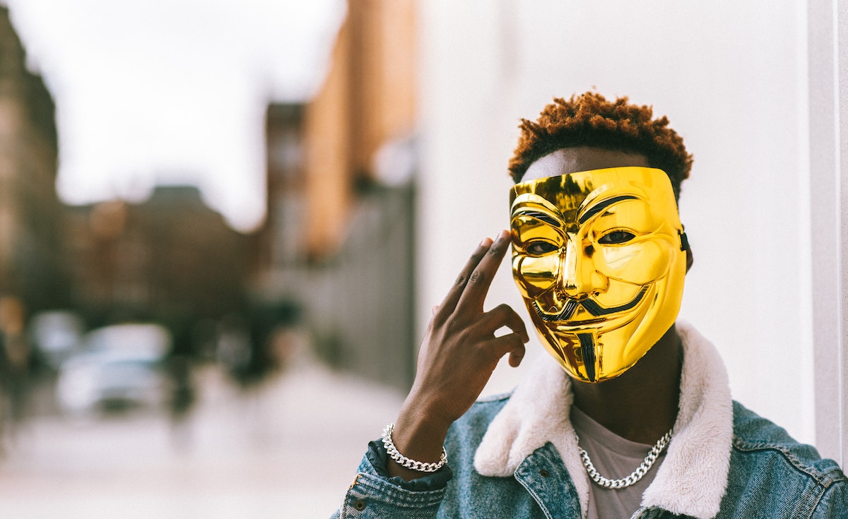 A person with a gold Guy Fawkes mask covering their face, wearing a denim jacket with a white fleece collar and two silver chain necklaces. Their dark-skinned hand is raised, touching the side of the mask, and their reddish-brown curly hair is visible above the mask. The background is blurred, showing light-colored buildings.