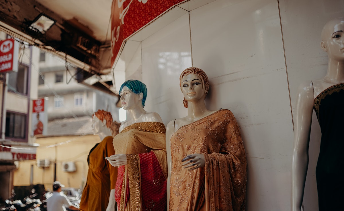 A line of four female mannequins, dressed in colorful saris, stand outside a clothing store. The mannequin in the foreground has light brown hair and a golden sari, while the one behind it has blue hair and a similar sari. The street and other buildings are visible in the blurred background.