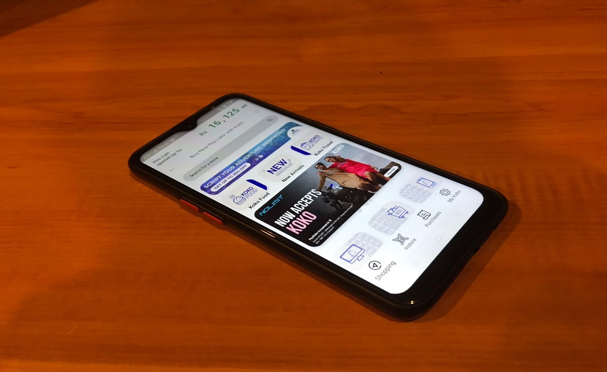 A black smartphone displaying the KOKO app interface, showing a balance of "Rs. 16,125" and various sections like "KOKO Food," "KOKO Travel," and a prominent banner "NOW ACCEPTS KOKO." The phone is lying diagonally on a wooden table.