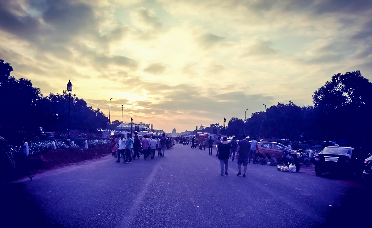 A wide-angle, low-light shot of a bustling street scene at dusk, likely in India, with numerous people walking along a purple-hued road. Trees line both sides, and distant buildings or monuments are silhouetted against a dramatic cloudy sky with hints of orange and yellow.