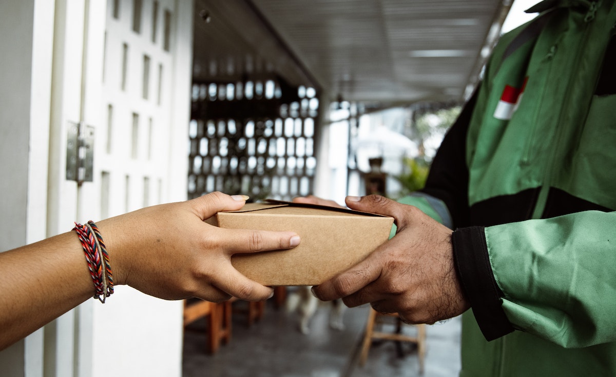 Close-up shot of two pairs of hands exchanging a small, brown cardboard box. On the left, a person with a multi-colored braided bracelet on their wrist extends their hands to receive the package. On the right, a person wearing a green delivery uniform with a red and white flag patch (possibly Indonesia) on the sleeve hands over the package. The background shows a blurry outdoor scene with white doors and some wooden furniture.