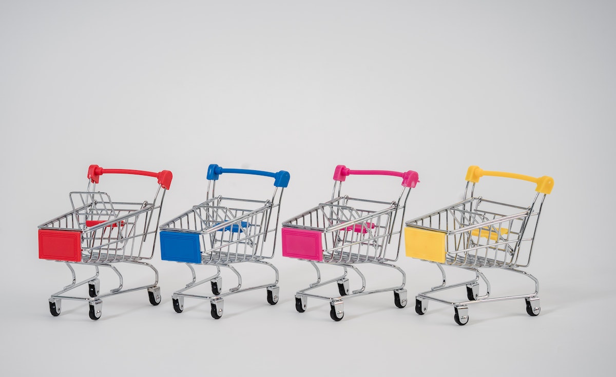 Four miniature shopping carts, each with a different colored handle and plastic base, are lined up in a row on a plain white background. From left to right, the colors are red, blue, pink, and yellow. The carts are facing slightly to the right.