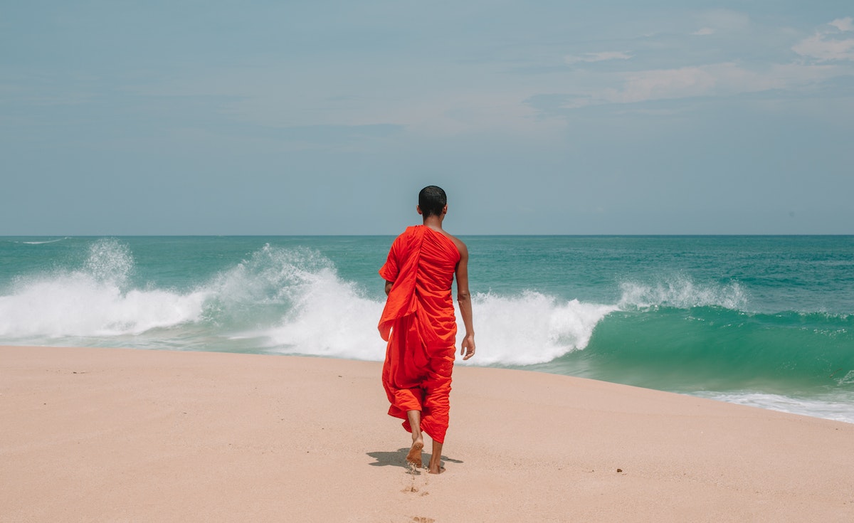 A person with a shaved head, wearing a bright orange-red monastic robe, walks barefoot on a sandy beach towards the ocean. Their back is to the viewer. Large waves crash in the turquoise-blue sea under a clear, light blue sky. Footprints are visible in the sand behind them.
