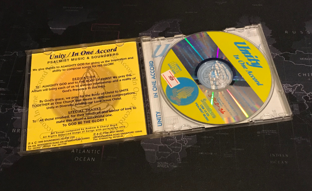 A CD case, open and displaying a colorful CD, is placed on a dark, possibly fabric, surface that appears to be a world map. The CD, titled "Unity In One Accord," has a bright yellow and blue design with text. The CD's liner notes, with acknowledgements and dedications, are visible on the left side of the open case. The map beneath shows outlines of continents and countries, with text like "ATLANTIC OCEAN," "SOUTH AFRICA," and "RUSSIA" partially visible, indicating a global context for the scene. The lighting suggests an indoor setting with some shadows.