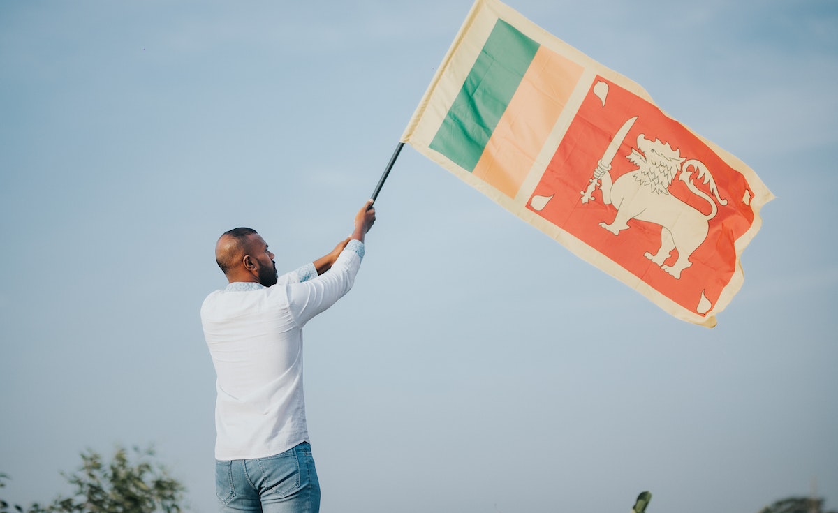 A man, seen from behind, stands holding the national flag of Sri Lanka aloft against a clear, light blue sky. He is wearing a white long-sleeved shirt and blue jeans. The flag, predominantly red with a golden lion holding a sword, and green and orange vertical stripes on the hoist side, is unfurled and waving. His hands are raised, firmly gripping the flag pole, suggesting a gesture of patriotism or support.