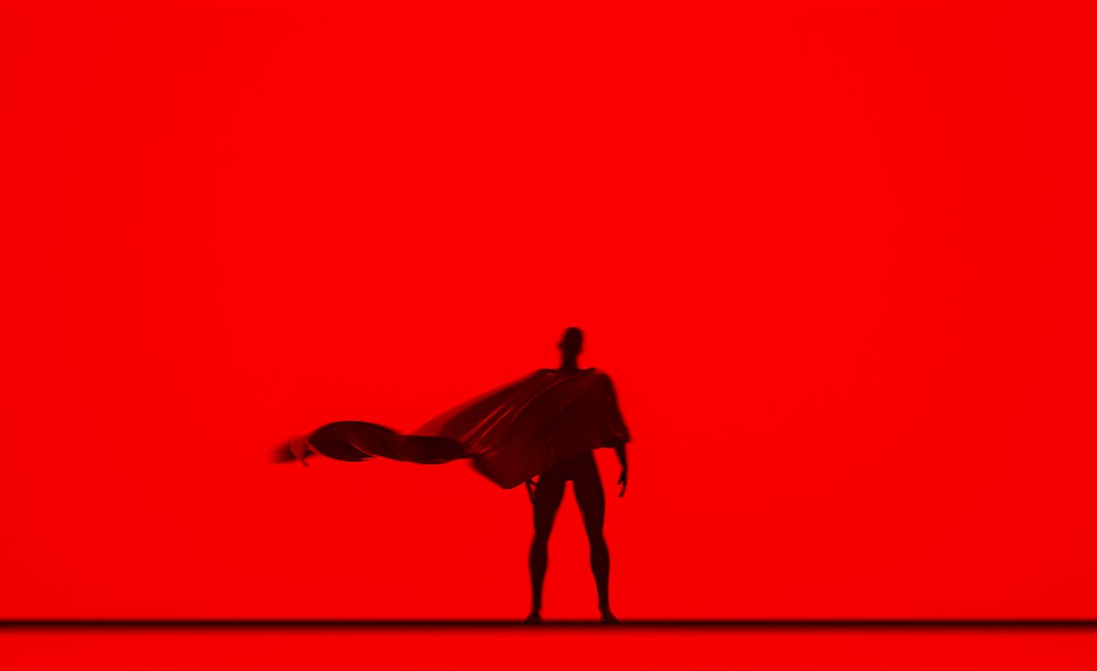 A silhouette of a figure, likely a superhero, stands against a solid, vibrant red background. The figure is facing forward with a cape dramatically flowing out to the left, suggesting movement or a powerful stance. There's a subtle horizontal line at the bottom, creating a ground plane for the figure. The overall image is striking due to its minimalist composition and strong color contrast.