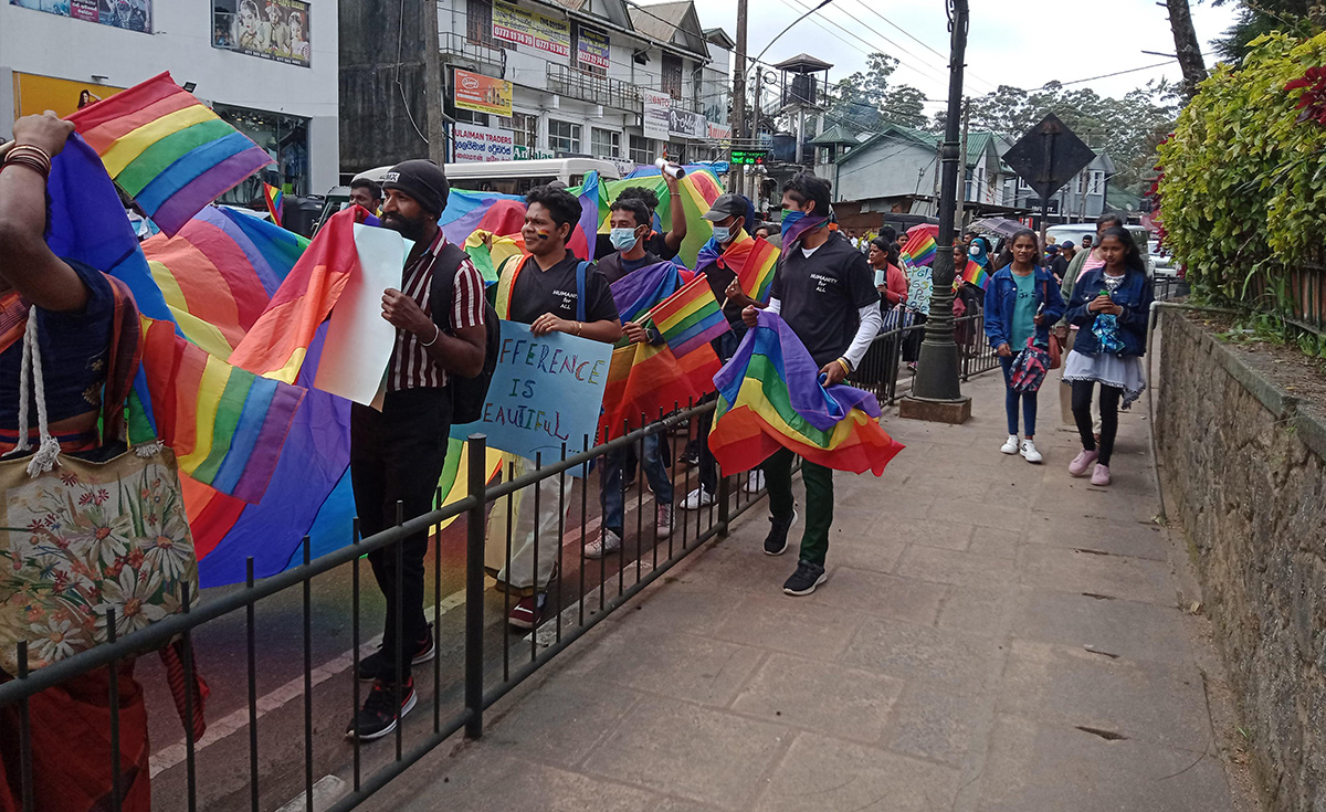 A group of people are walking on a paved pathway next to a street, holding rainbow flags and signs. The setting appears to be an urban or semi-urban area with buildings and some greenery visible in the background. Many individuals in the foreground are carrying rainbow flags, some draped over their shoulders, while others hold them up. One person on the left is holding a large rainbow flag with both hands. Another person in the center is holding a white sign. Further along the path, other participants are visible, some wearing face masks. The overall atmosphere suggests a parade or a public demonstration.