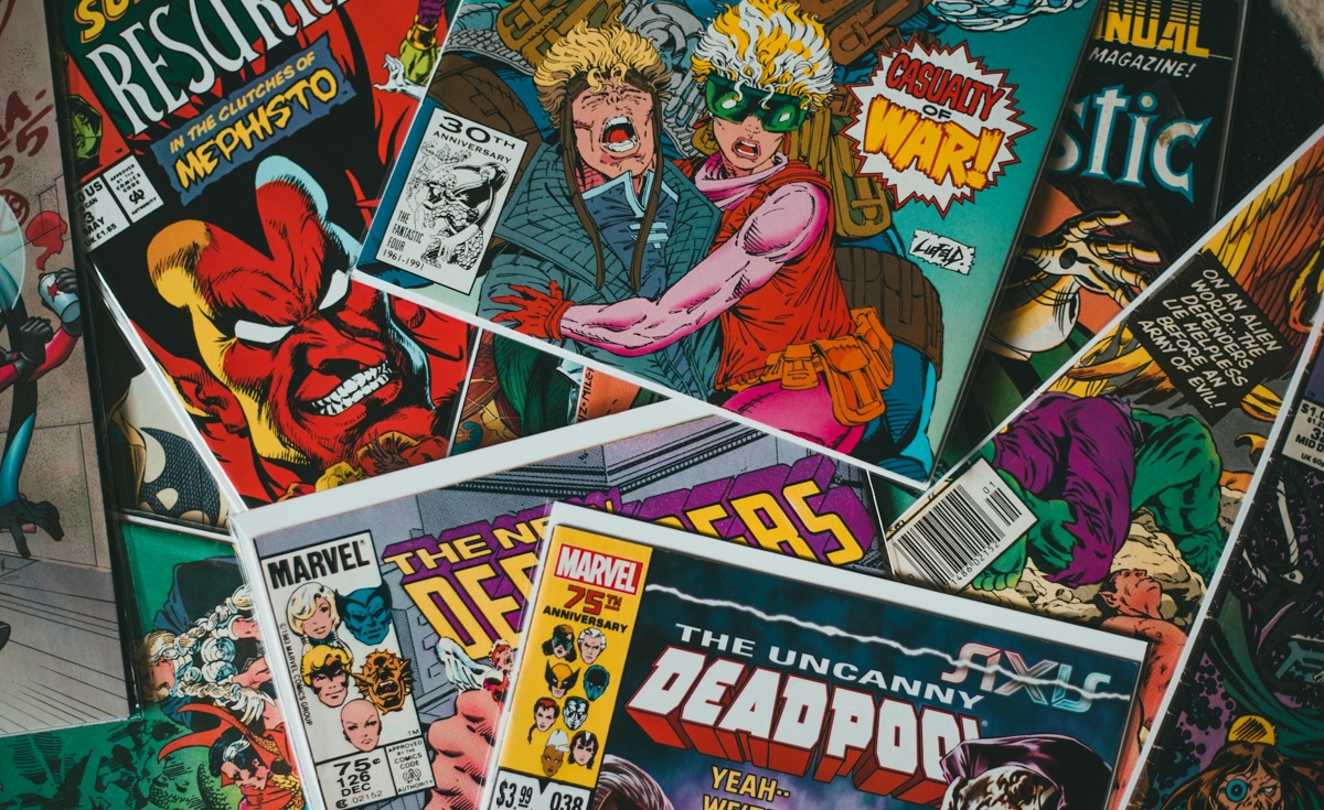 A close-up, overhead shot displays a collection of comic books, predominantly from Marvel Comics, spread out and overlapping. The vibrant and dynamic artwork from the covers is the main focus. Some identifiable titles and characters include: * A comic with a large red devil-like character on the left, possibly "Mephisto." * A comic featuring two characters, one with blonde spiky hair and the other with yellow hair and green glasses, with "CASUALTY OF WAR!" prominently displayed. * Another comic shows "THE UNCANNY DEADPOOL" with a "Marvel 75th Anniversary" logo. * Various other Marvel logos and character art are visible on partially obscured covers. The overall impression is a colorful and detailed jumble of classic comic book art.